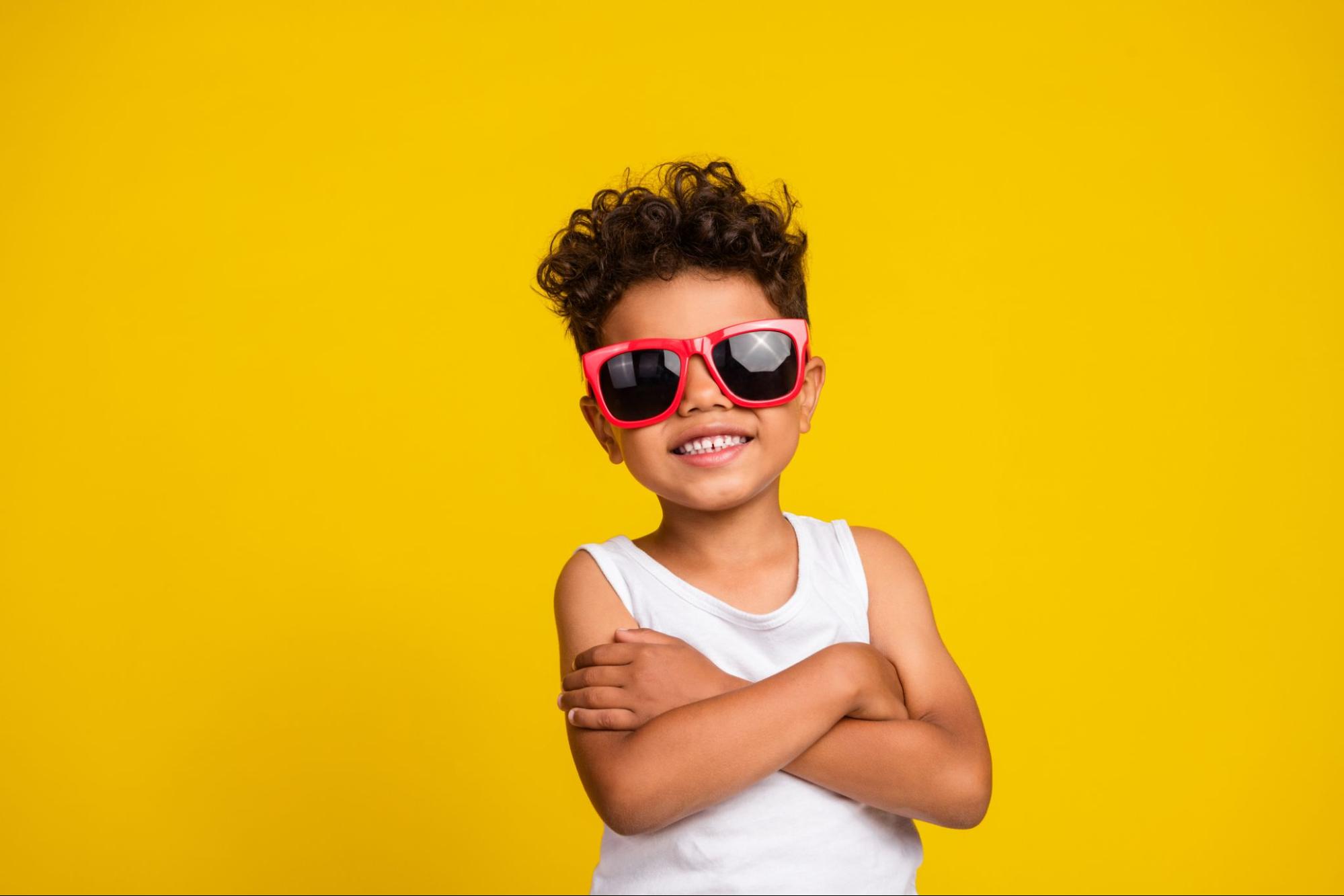 Young boy wearing red kids sunglasses with arms crossed against a yellow background