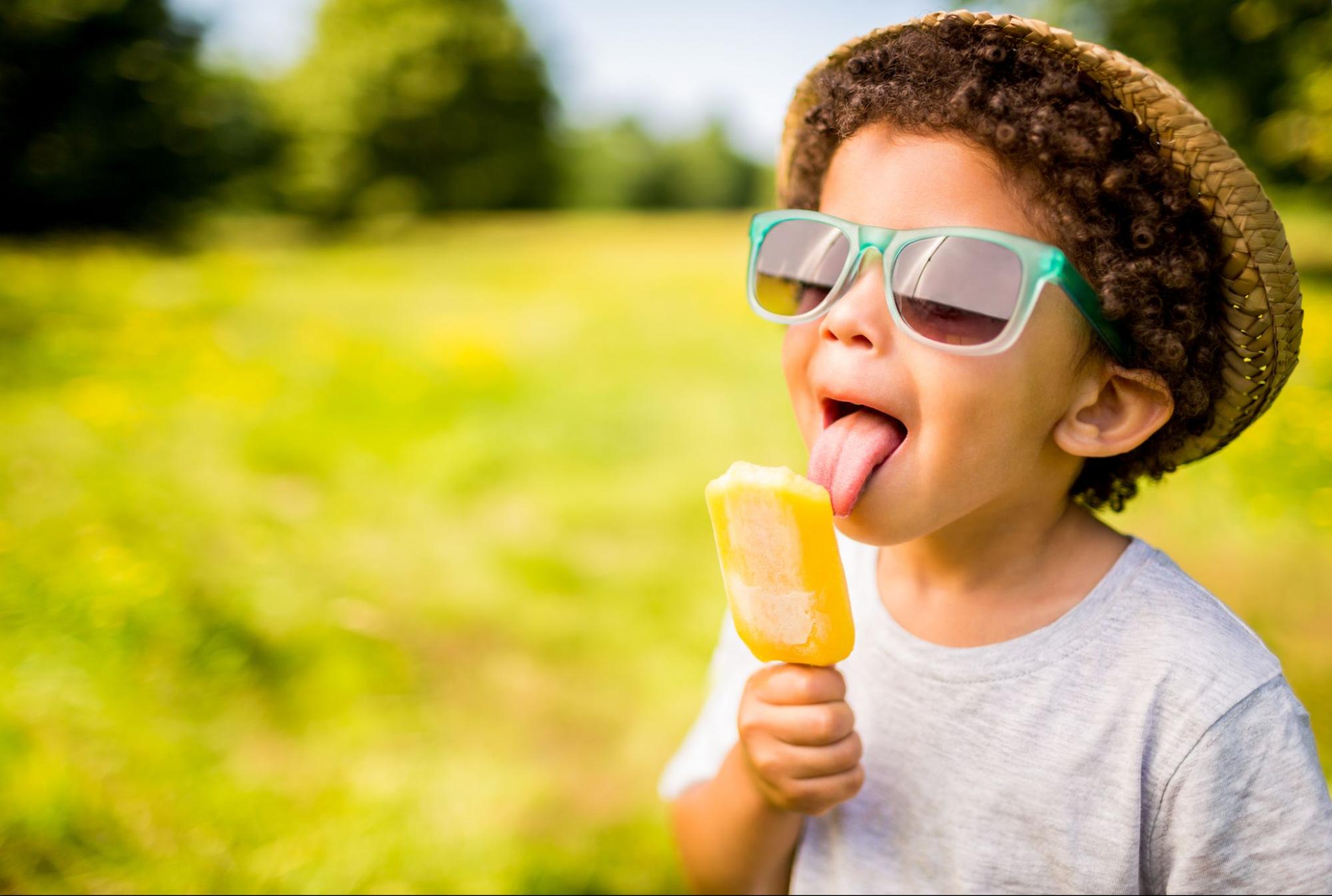 Young child in teal sunglasses and straw hat eating a popsicle outside on a sunny day