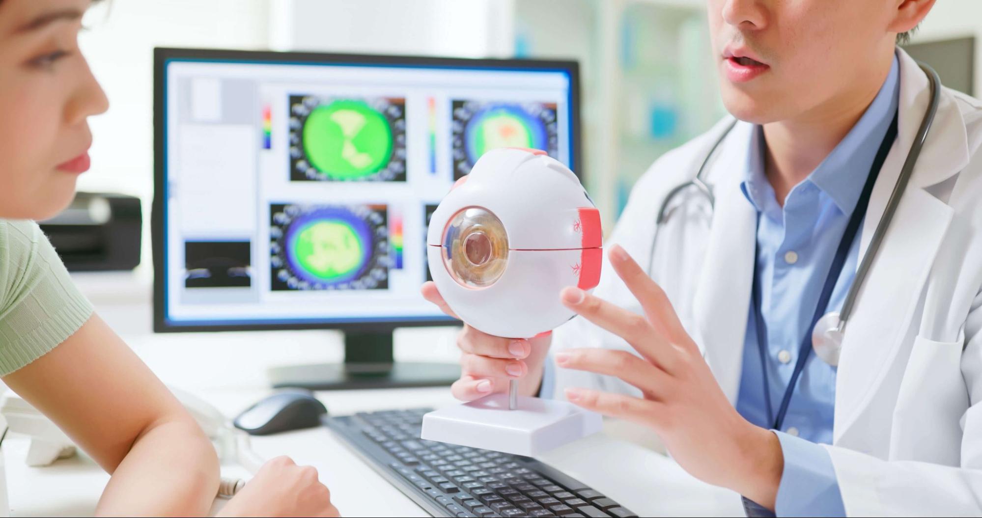Eye doctor explaining astigmatism to a patient using an eye model with corneal topography scans on screen