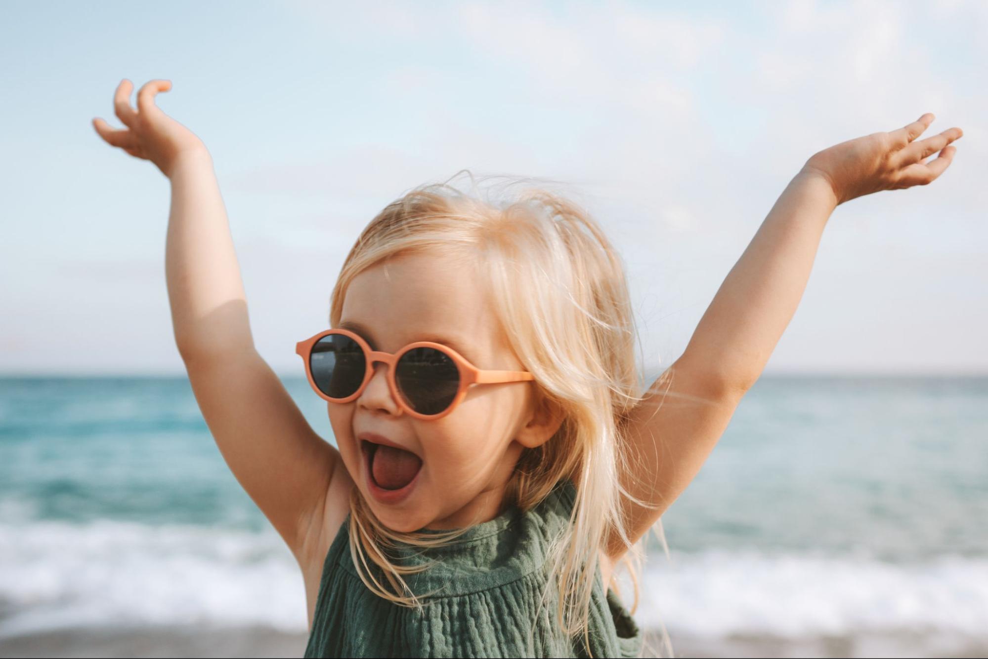 Happy toddler wearing orange round sunglasses at the beach with arms raised