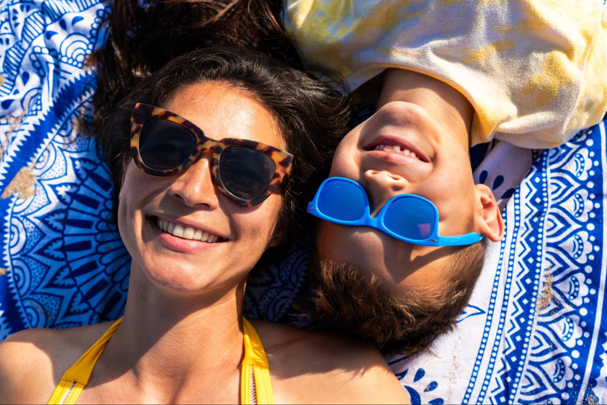 Mother and child lying on a beach blanket showing the difference between adult and kids sunglasses side by side