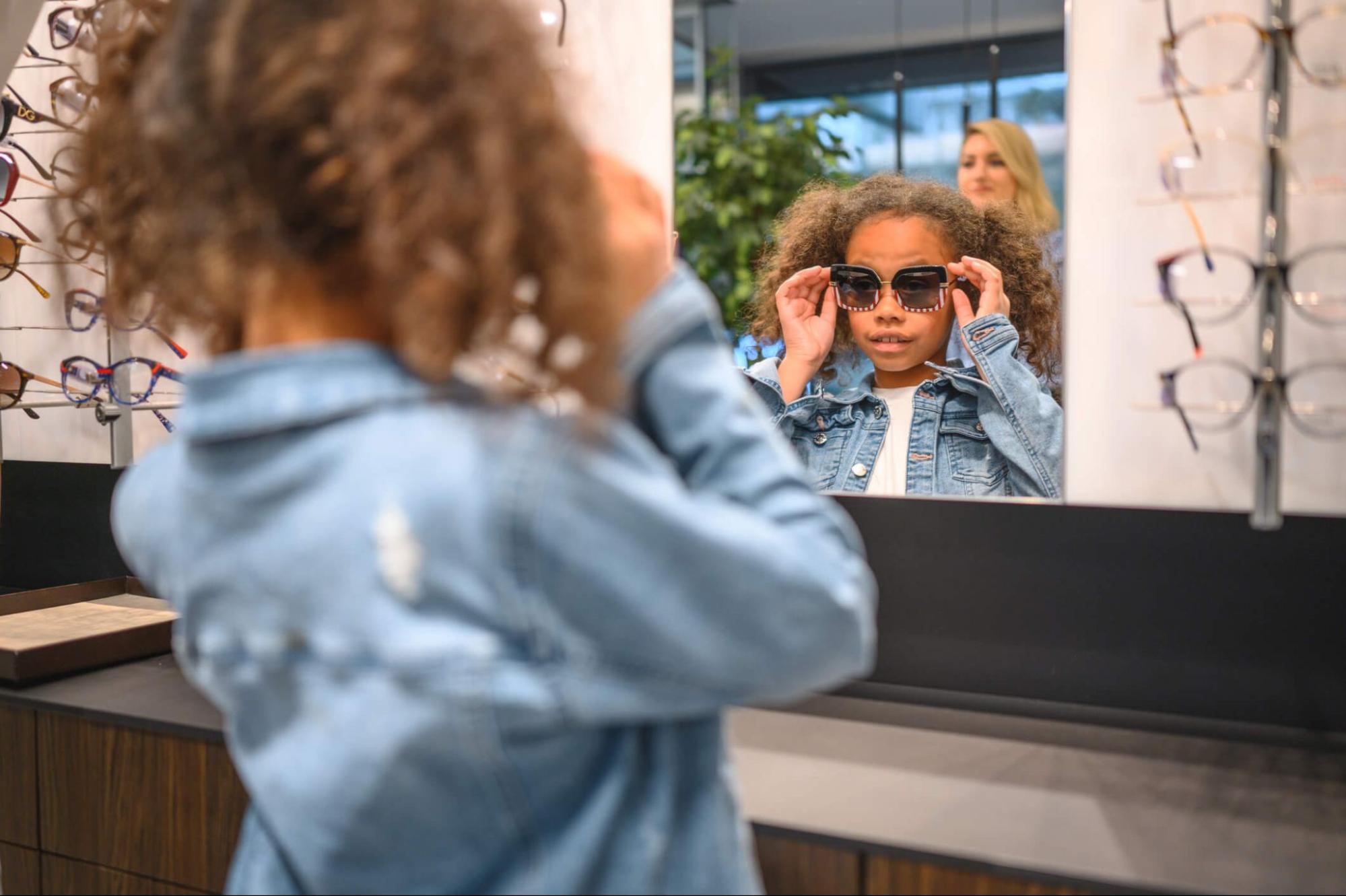 Young girl trying on children's sunglasses UV protection styles in a store mirror